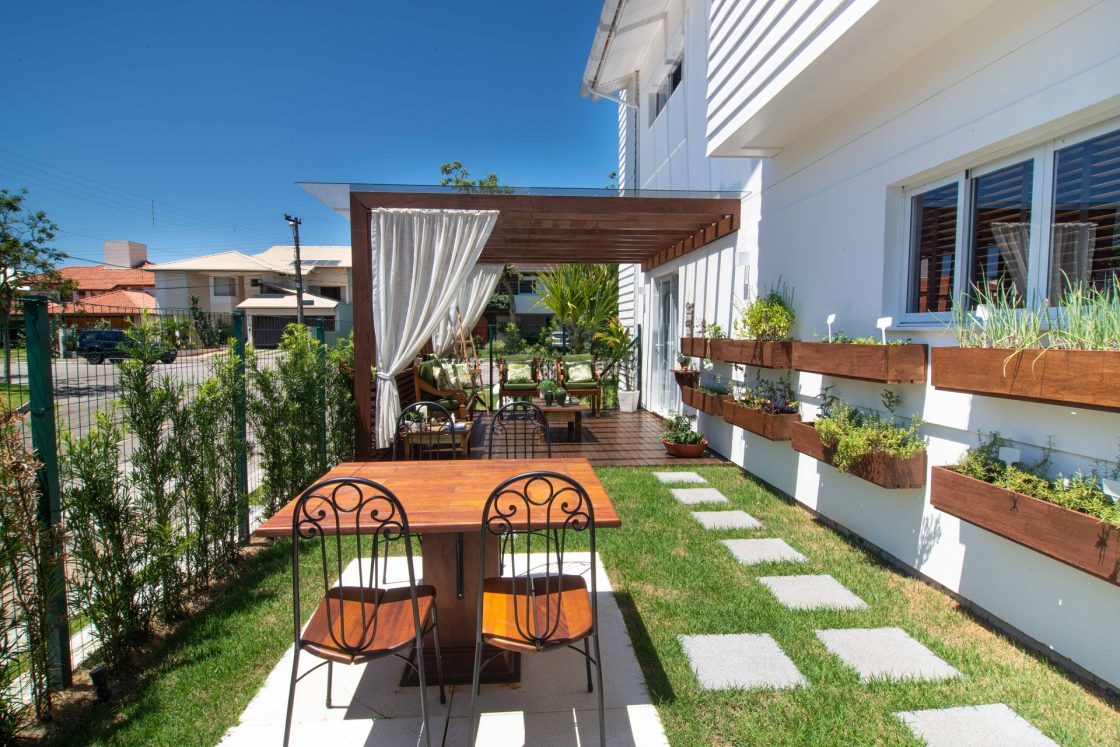 Jardim externo residencial com plantas em floreiras na parede, área de lazer com mesa de madeira e cadeiras, pergolado com cortinas brancas e caminho de pedras no gramado