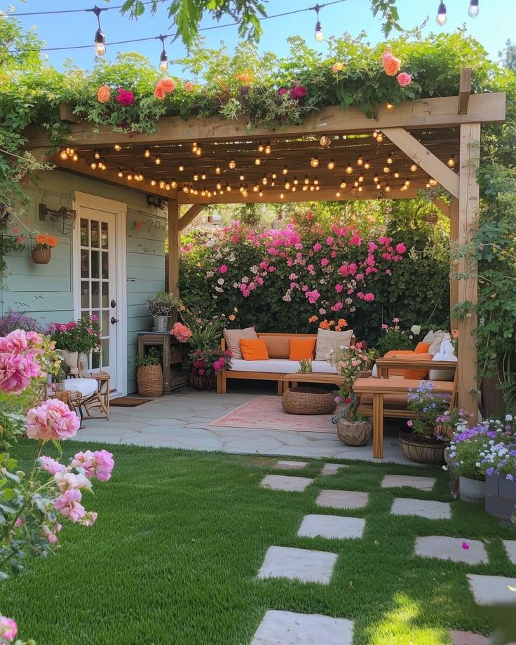 Espaço de jardim com pergolado de madeira, sofá com almofadas laranjas, luzes decorativas e muitas flores coloridas